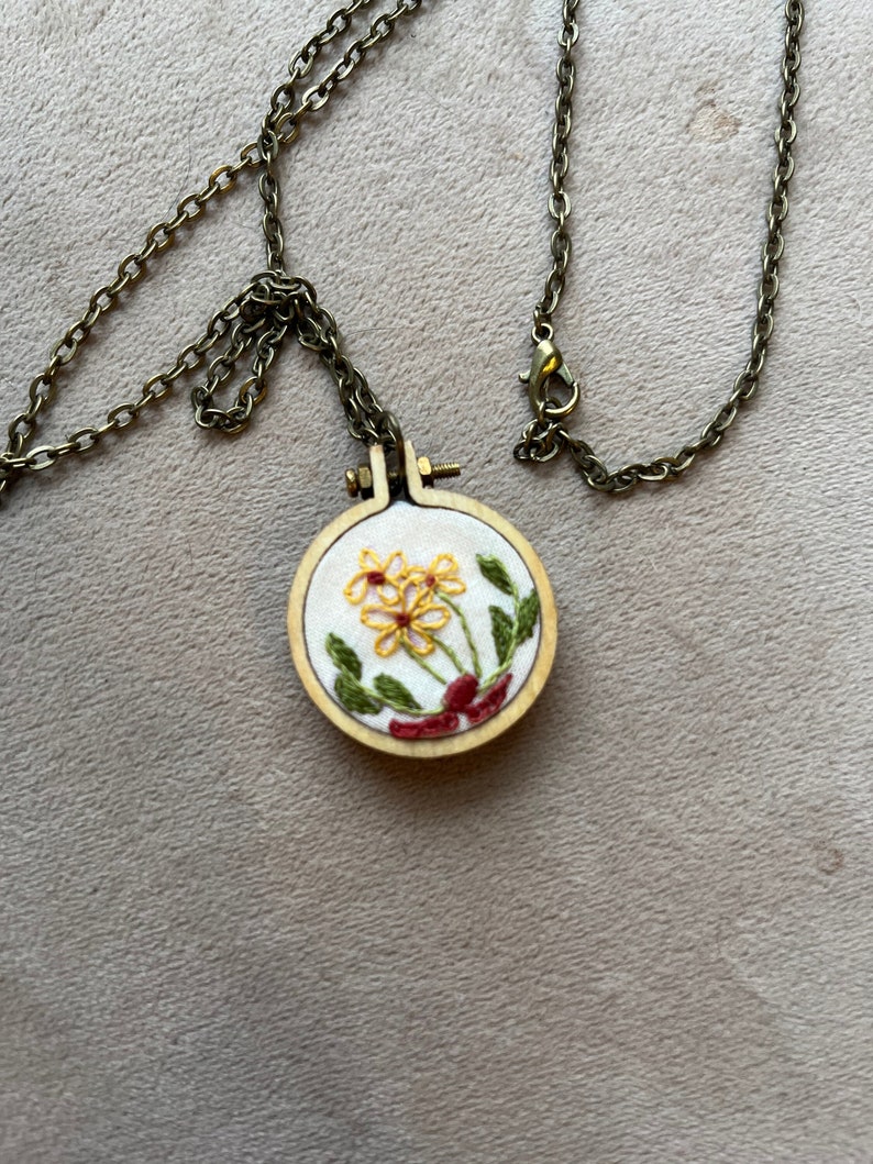 Pu&ograve; includere: Una delicata collana a catena dorata con un piccolo ciondolo rotondo in legno con fiori gialli ricamati con foglie verdi e steli rossi.