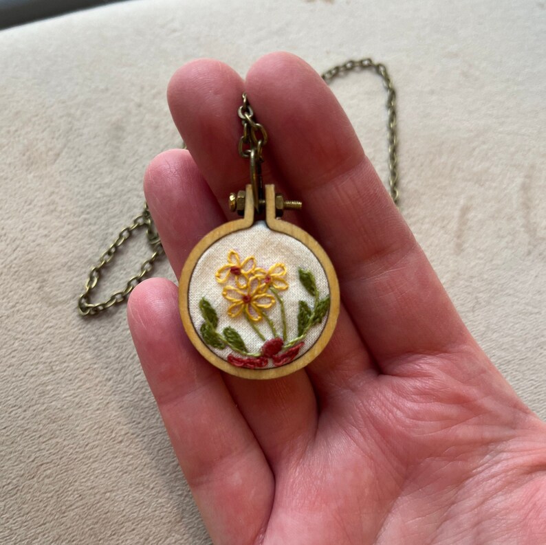 Pu&ograve; includere: Una collana delicata con un piccolo ciondolo rotondo in legno con fiori gialli ricamati con foglie verdi su sfondo bianco.