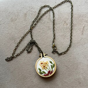 Pu&ograve; includere: Una delicata collana a catena dorata con un piccolo ciondolo rotondo in legno con fiori gialli ricamati con foglie verdi e accenti rossi.
