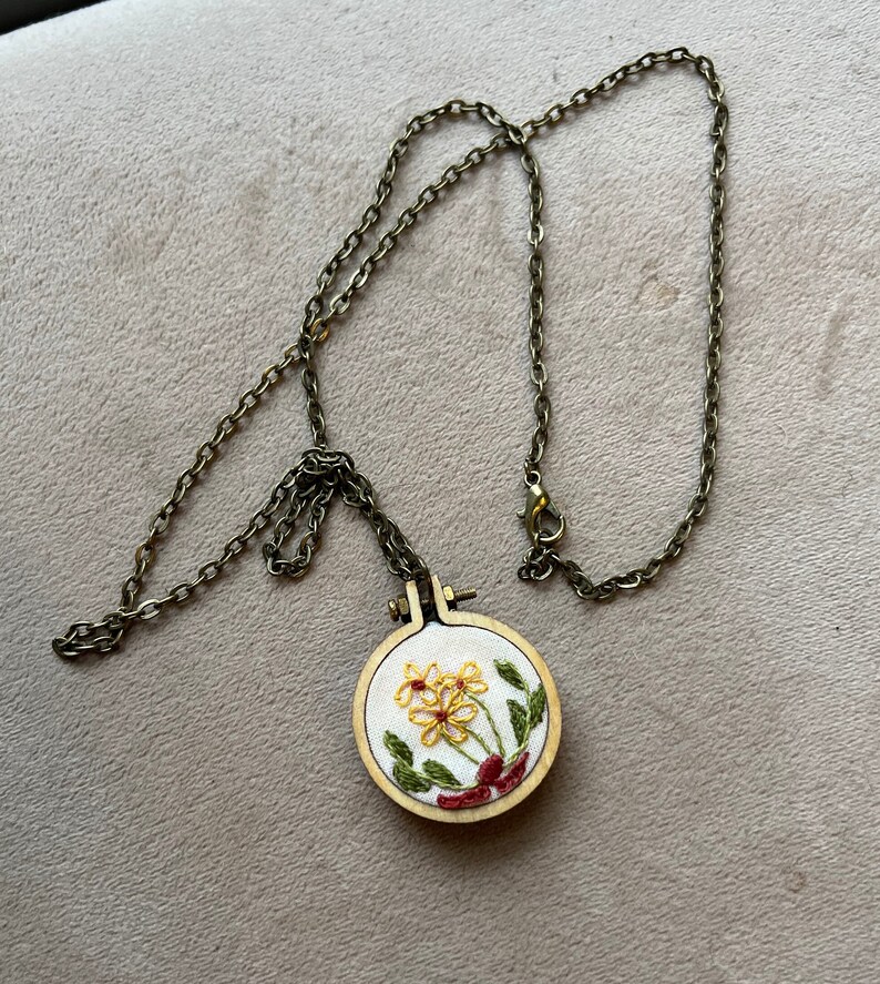 Pu&ograve; includere: Una delicata collana a catena dorata con un piccolo ciondolo rotondo in legno con fiori gialli ricamati con foglie verdi e accenti rossi.