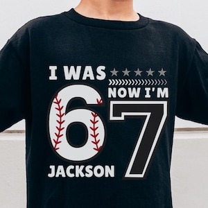 May include: Black t-shirt with the text "I WAS now I'M 67 JACKSON" and a baseball graphic. The shirt is worn by a child wearing sunglasses and a baseball cap. The child is holding up two fingers on each hand.