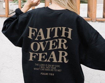 Może przedstawiać: Czarny sweter z nadrukiem "FAITH OVER FEAR" w brązowych literach. Tekst "THE LORD IS ON MY SIDE: I WILL NOT FEAR WHAT CAN MAN DO TO ME? PSALMS 118:6" również jest nadrukowany w brązowych literach.