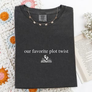 May include: A charcoal gray short-sleeved t-shirt featuring the white text "our favorite plot twist" centered above a graphic of an open book with two small baby footprints emerging from the pages, printed on a soft cotton fabric.