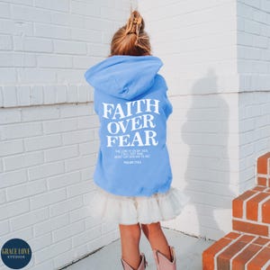 Puede incluir: Una sudadera con capucha azul claro con el texto "FAITH OVER FEAR" en blanco. La sudadera tambi&eacute;n incluye el texto "THE LORD IS ON MY SIDE, I WILL NOT FEAR, WHAT CAN MAN DO TO ME? PSALMS 118:6" en blanco m&aacute;s peque&ntilde;o.