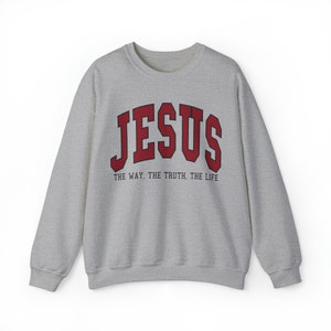 Może przedstawiać: Szara bluza z czerwonym nadrukiem "JESUS" w stylu uniwersyteckim. Pod nadrukiem znajduje się tekst "THE WAY. THE TRUTH. THE LIFE".