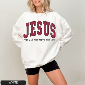 Może przedstawiać: Biała bluza z czerwonym nadrukiem "JESUS" i tekstem "THE WAY, THE TRUTH, THE LIFE" w kolorze czarnym.