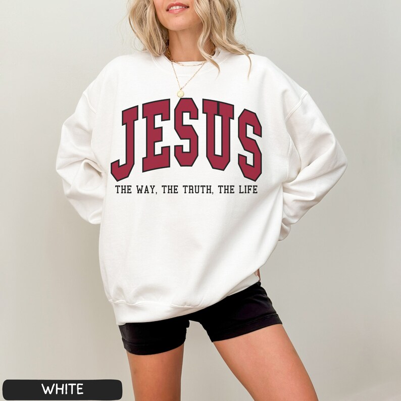 Może przedstawiać: Biała bluza z czerwonym nadrukiem "JESUS" i tekstem "THE WAY, THE TRUTH, THE LIFE" w kolorze czarnym.