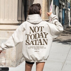 May include: A beige hoodie with the text "NOT TODAY SATAN" in brown lettering. The text "THEREFORE SUBMIT TO GOD. RESIST THE DEVIL AND HE WILL FLEE FROM YOU. JAMES 4:7" is printed in smaller brown lettering below the main text.