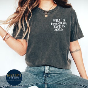 Peut inclure: Un t-shirt gris fonc&eacute; avec le texte "WHAT A FRIEND WE HAVE IN JESUS" imprim&eacute; en blanc sur le devant. Le t-shirt est port&eacute; par une personne aux cheveux longs bruns et un collier en or. La personne porte un jean bleu.