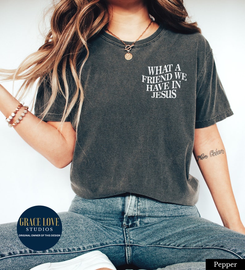 Peut inclure: Un t-shirt gris fonc&eacute; avec le texte "WHAT A FRIEND WE HAVE IN JESUS" imprim&eacute; en blanc sur le devant. Le t-shirt est port&eacute; par une personne aux cheveux longs bruns et un collier en or. La personne porte un jean bleu.