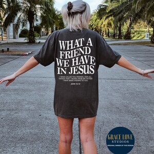 Peut inclure: Un t-shirt noir avec du texte blanc qui dit "What a friend we have in Jesus." Le texte est en gras, avec une police de caract&egrave;res en blocs. Le t-shirt comprend &eacute;galement un verset biblique de Jean 15:14. Le t-shirt est port&eacute; par une personne sur un parking.