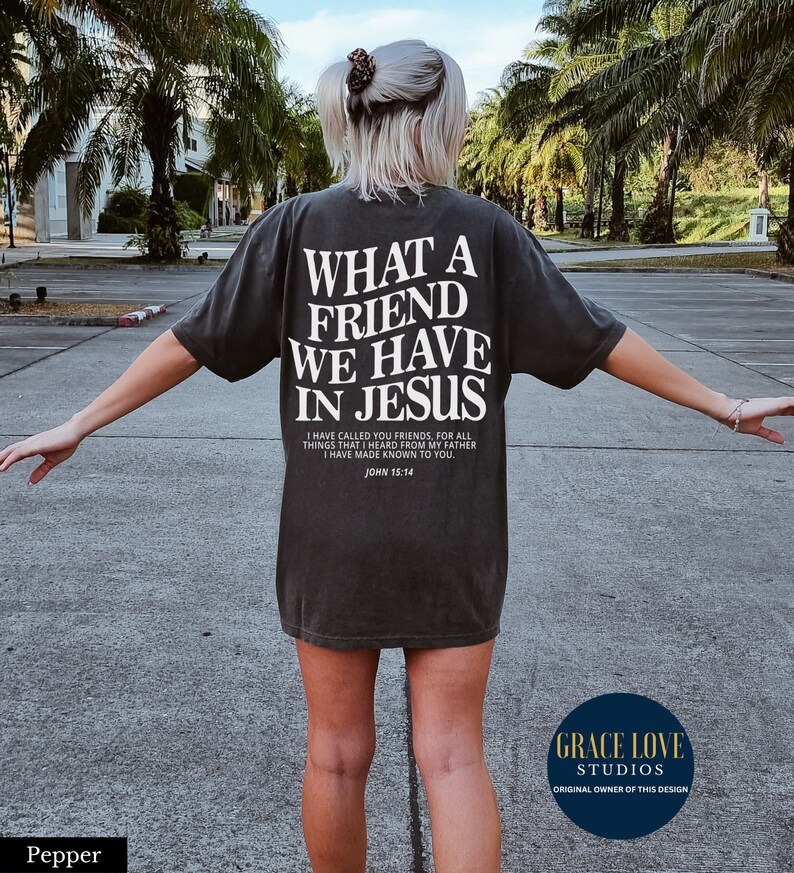 Peut inclure: Un t-shirt noir avec du texte blanc qui dit "What a friend we have in Jesus." Le texte est en gras, avec une police de caract&egrave;res en blocs. Le t-shirt comprend &eacute;galement un verset biblique de Jean 15:14. Le t-shirt est port&eacute; par une personne sur un parking.