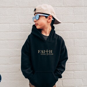 Puede incluir: Sudadera con capucha negra con el texto "FAITH OVER FEAR" y un gr&aacute;fico de cruz en dorado. El modelo lleva una gorra de b&eacute;isbol beige y gafas de sol azules.