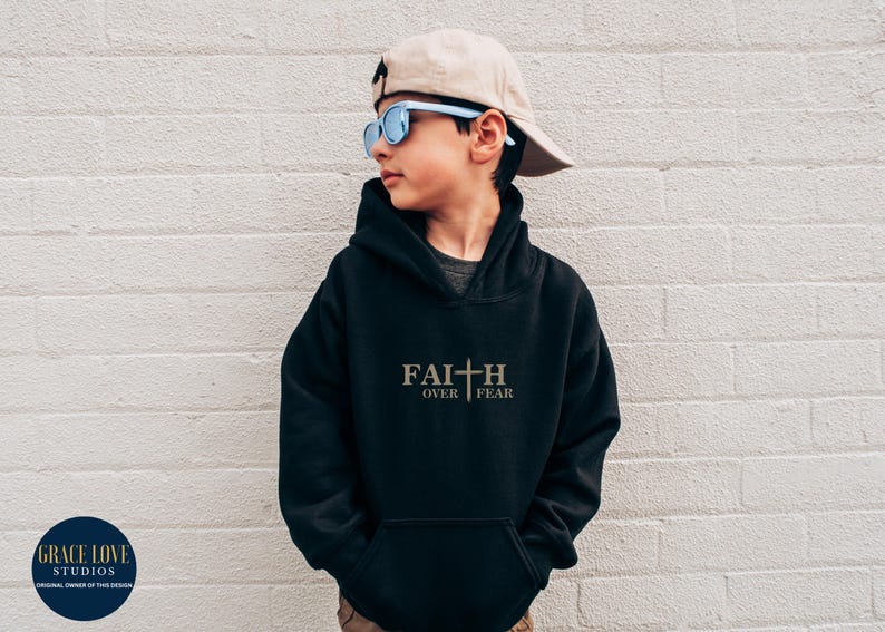 Puede incluir: Sudadera con capucha negra con el texto "FAITH OVER FEAR" y un gr&aacute;fico de cruz en dorado. El modelo lleva una gorra de b&eacute;isbol beige y gafas de sol azules.