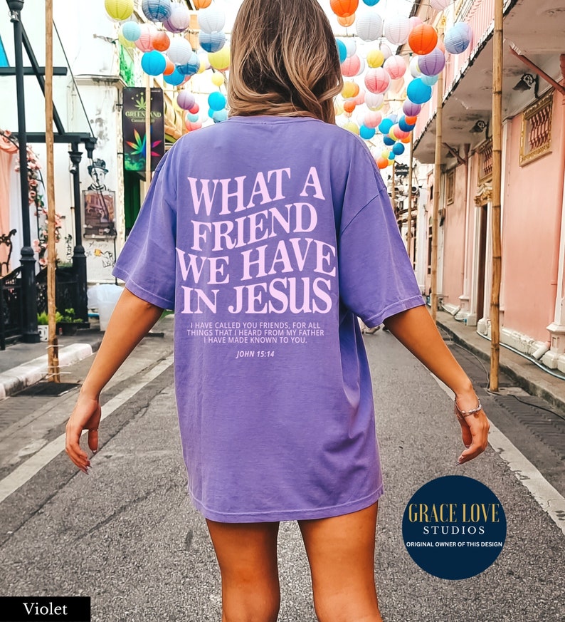 Peut inclure: Un t-shirt violet avec le texte "What a Friend We Have in Jesus" en lettres roses. Le t-shirt comprend &eacute;galement le verset biblique Jean 15:14 en lettres roses plus petites. Le t-shirt est port&eacute; par une personne qui marche dans la rue.