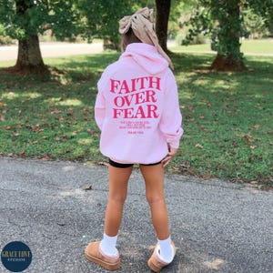Puede incluir: Sudadera rosa con las palabras "Faith Over Fear" en letras rojas en la parte posterior. La sudadera tambi&eacute;n tiene una cita b&iacute;blica de los Salmos 118:6.