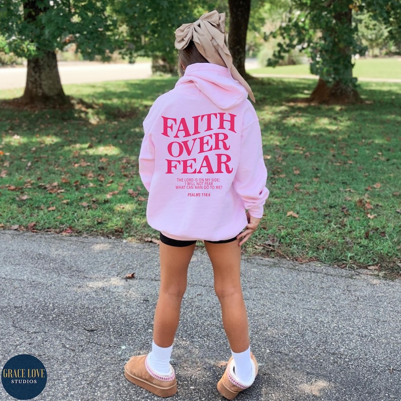 Puede incluir: Sudadera rosa con las palabras "Faith Over Fear" en letras rojas en la parte posterior. La sudadera tambi&eacute;n tiene una cita b&iacute;blica de los Salmos 118:6.