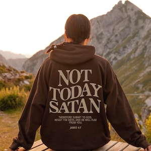 May include: A brown hoodie with the text "NOT TODAY SATAN" in beige lettering. The hoodie also features the text "THEREFORE SUBMIT TO GOD. RESIST THE DEVIL AND HE WILL FLEE FROM YOU. JAMES 4:7" in smaller beige lettering.