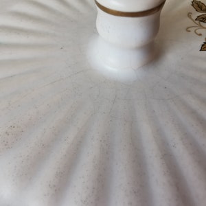 May include: Close-up of a white ceramic lid with a gold-trimmed handle. The lid has a ridged pattern and is decorated with black and gold floral accents. The ceramic has visible cracks.