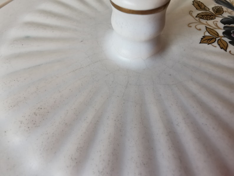 May include: Close-up of a white ceramic lid with a gold-trimmed handle. The lid has a ridged pattern and is decorated with black and gold floral accents. The ceramic has visible cracks.