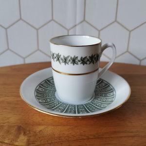 Op de afbeelding: Witte theekop en schotel set met gouden rand. De theekop heeft een groen bladerenpatroon en een gouden band. De schotel heeft een bijpassend groen en wit ontwerp. De set staat op een houten oppervlak.