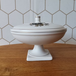 May include: A white ceramic pedestal dish with a lid. The dish has a round bowl shape and sits on a square base. The lid is decorated with a floral design and gold trim. The dish is likely vintage.