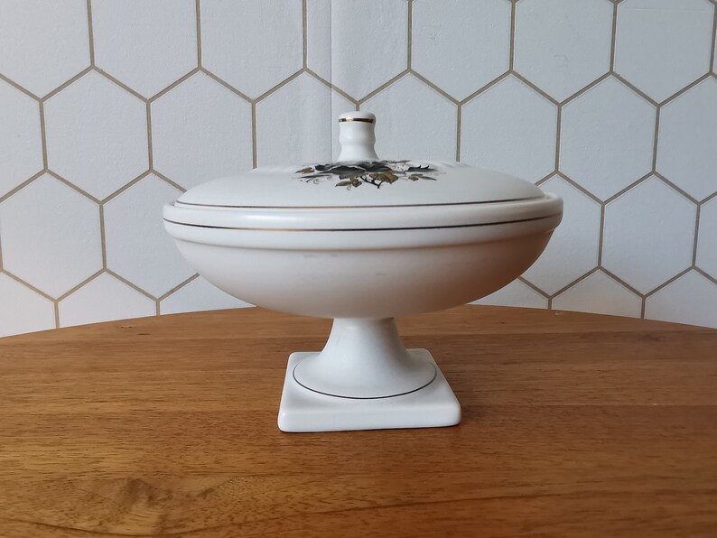 May include: A white ceramic pedestal dish with a lid. The dish has a round bowl shape and sits on a square base. The lid is decorated with a floral design and gold trim. The dish is likely vintage.