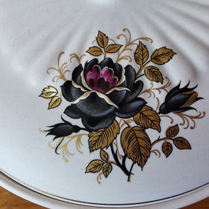 May include: White ceramic serving dish lid with a black rose and gold leaf design. The rose has a deep purple center. The lid has a scalloped edge and a gold trim. A decorative piece for the kitchen.