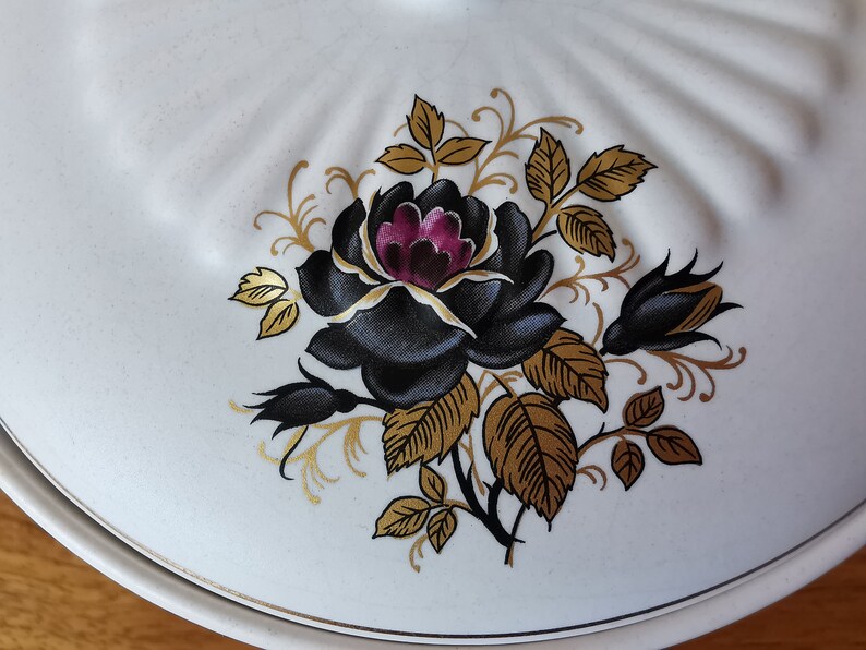 May include: White ceramic serving dish lid with a black rose and gold leaf design. The rose has a deep purple center. The lid has a scalloped edge and a gold trim. A decorative piece for the kitchen.