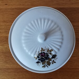 May include: A white ceramic serving dish with a lid. The lid features a raised, fan-like design and a small knob. A floral motif with black and purple roses and gold accents decorates the dish.