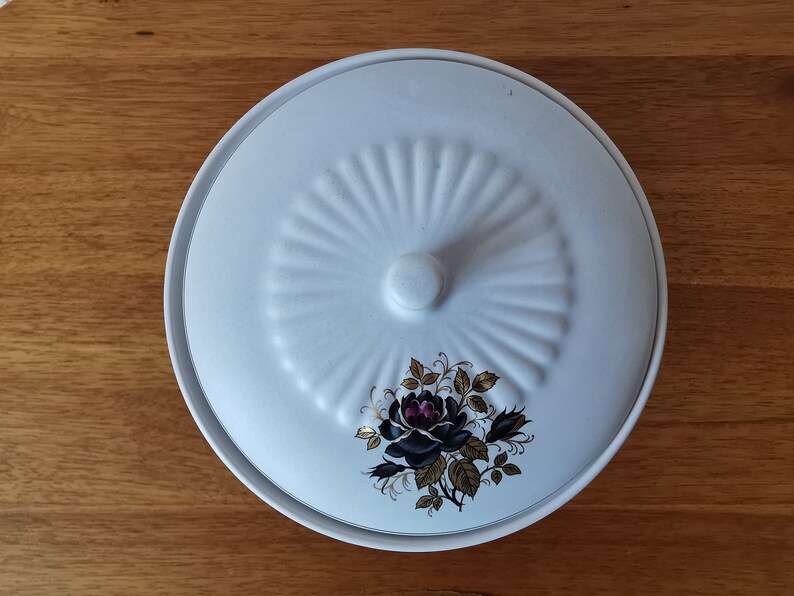 May include: A white ceramic serving dish with a lid. The lid features a raised, fan-like design and a small knob. A floral motif with black and purple roses and gold accents decorates the dish.