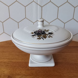 May include: White ceramic lidded serving dish with a pedestal base. The lid features a black and purple rose design with gold accents. The dish has gold trim and a small finial on top. A decorative piece for the home.