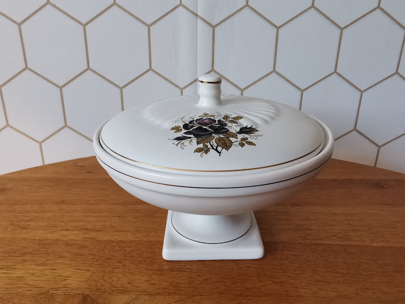 May include: White ceramic lidded serving dish with a pedestal base. The lid features a black and purple rose design with gold accents. The dish has gold trim and a small finial on top. A decorative piece for the home.