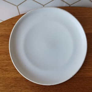 May include: A round, white ceramic plate with a slightly raised edge. The plate is set on a wooden surface, with a white and gray geometric tile background visible in the upper left corner. This plate is suitable for serving food.
