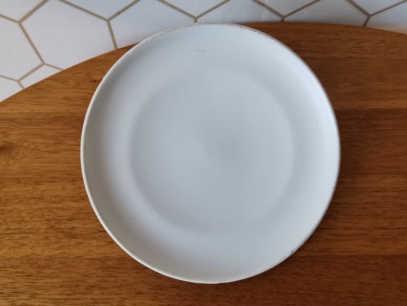 May include: A round, white ceramic plate with a slightly raised edge. The plate is set on a wooden surface, with a white and gray geometric tile background visible in the upper left corner. This plate is suitable for serving food.