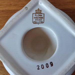 May include: A white ceramic object with a circular opening and a square base. The object has a gold emblem that reads "FLORA KERAMIEK GOUDA HOLLAND". The number "2009" is printed in red.