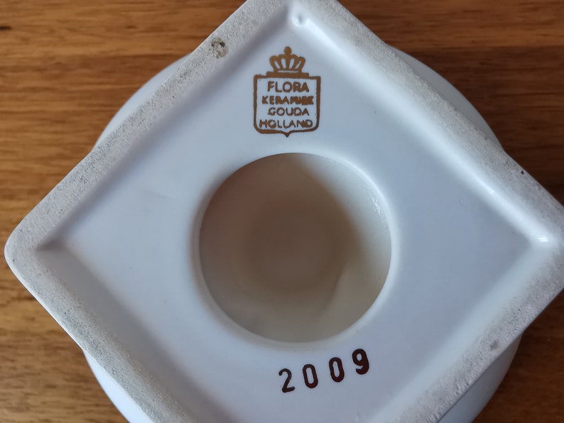 May include: A white ceramic object with a circular opening and a square base. The object has a gold emblem that reads "FLORA KERAMIEK GOUDA HOLLAND". The number "2009" is printed in red.