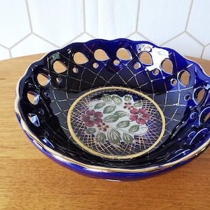 Puede incluir: Un cuenco de cerámica azul cobalto con borde festoneado y diseño calado. El cuenco presenta un motivo floral en el centro, con un borde dorado. El cuenco es probablemente para uso decorativo.