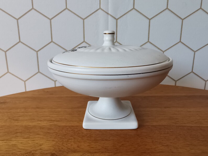 May include: White ceramic lidded serving dish with gold trim. The dish has a pedestal base and a lid with a small handle. The lid features a floral design. The dish is on a wooden surface.