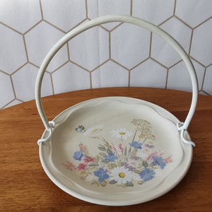 Puede incluir: Una fuente para servir de color crema con un asa blanca. El plato presenta un diseño floral con margaritas, flores azules y una mariposa. El asa está sujeta al plato con soportes metálicos blancos. El plato está sobre una superficie de madera.