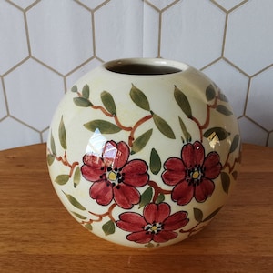 Op de afbeelding: Een ronde, crèmekleurige keramische vaas met een bloemenontwerp. De vaas heeft drie grote rode bloemen met donkere centra, groene bladeren en bruine stelen. De vaas staat op een houten oppervlak.