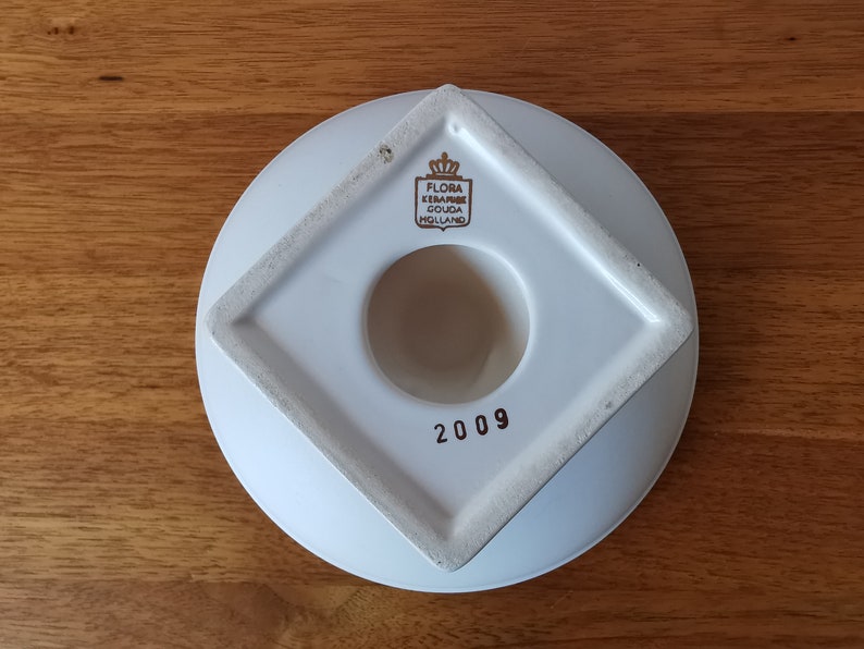 May include: A white ceramic wall pocket with a circular base and a square opening. The pocket has a diamond-shaped frame around the opening. The text "FLORA KERAMIEK GOUDA HOLLAND" and the number "2009" are visible.