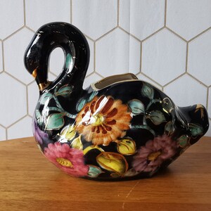 Pode incluir: Vaso de cerâmica preto em forma de cisne com desenhos florais em laranja, rosa e amarelo. O vaso tem um acabamento brilhante e detalhes dourados. O pescoço do cisne curva-se graciosamente e o corpo é decorado com folhas.