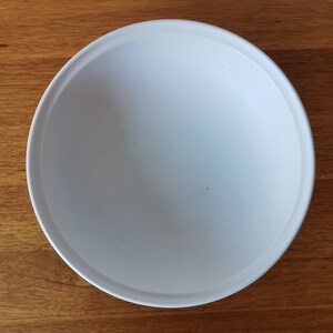 May include: A white, round ceramic plate with a slightly raised rim. The plate is centered on a wooden surface. The plate's interior is smooth and empty, ready for food. The plate is approximately 10 inches in diameter.
