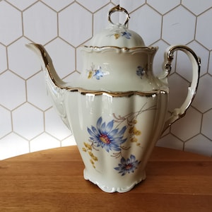 Può includere: Caffettiera in ceramica color crema con coperchio, con un motivo floreale con fiori blu e rossi e finiture dorate. Anche il manico e il beccuccio sono rifiniti in oro. Un pezzo decorativo per servire caffè o tè.