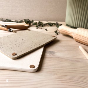May include: Two rectangular ceramic tiles with cork feet. The top tile is speckled with brown and beige. The bottom tile is white. The tiles are on a light wood surface.
