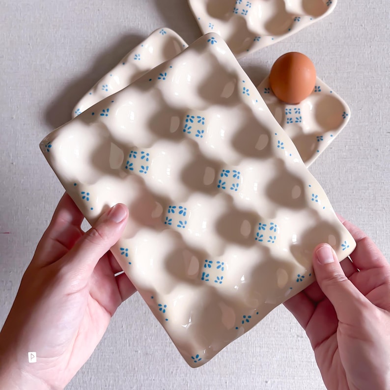 May include: A cream-coloured ceramic egg tray featuring a repeating pattern of small blue floral motifs. The rectangular tray has multiple rounded indentations for holding eggs, with one brown egg resting in a smaller matching tray nearby.