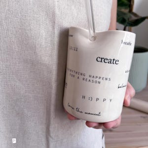 May include: A cream-colored ceramic tumbler with a wavy top edge and a clear glass straw. The surface features black printed text including "create," "11:11," "EVERYTHING HAPPENS FOR A REASON," "H :) P P Y," "love the moment," "breathe," and "balance."
