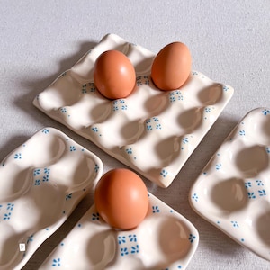 May include: Three cream-coloured ceramic egg trays with a glossy finish, each featuring a repeating pattern of small blue floral motifs. The trays have moulded indentations for eggs, and three brown chicken eggs are placed within the indentations.