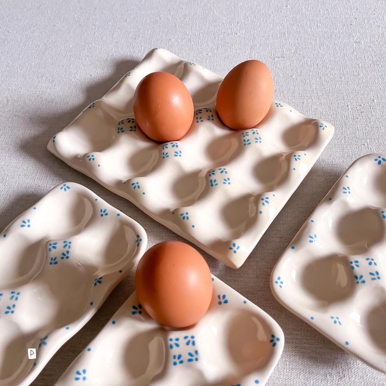 May include: Three cream-coloured ceramic egg trays with a glossy finish, each featuring a repeating pattern of small blue floral motifs. The trays have moulded indentations for eggs, and three brown chicken eggs are placed within the indentations.